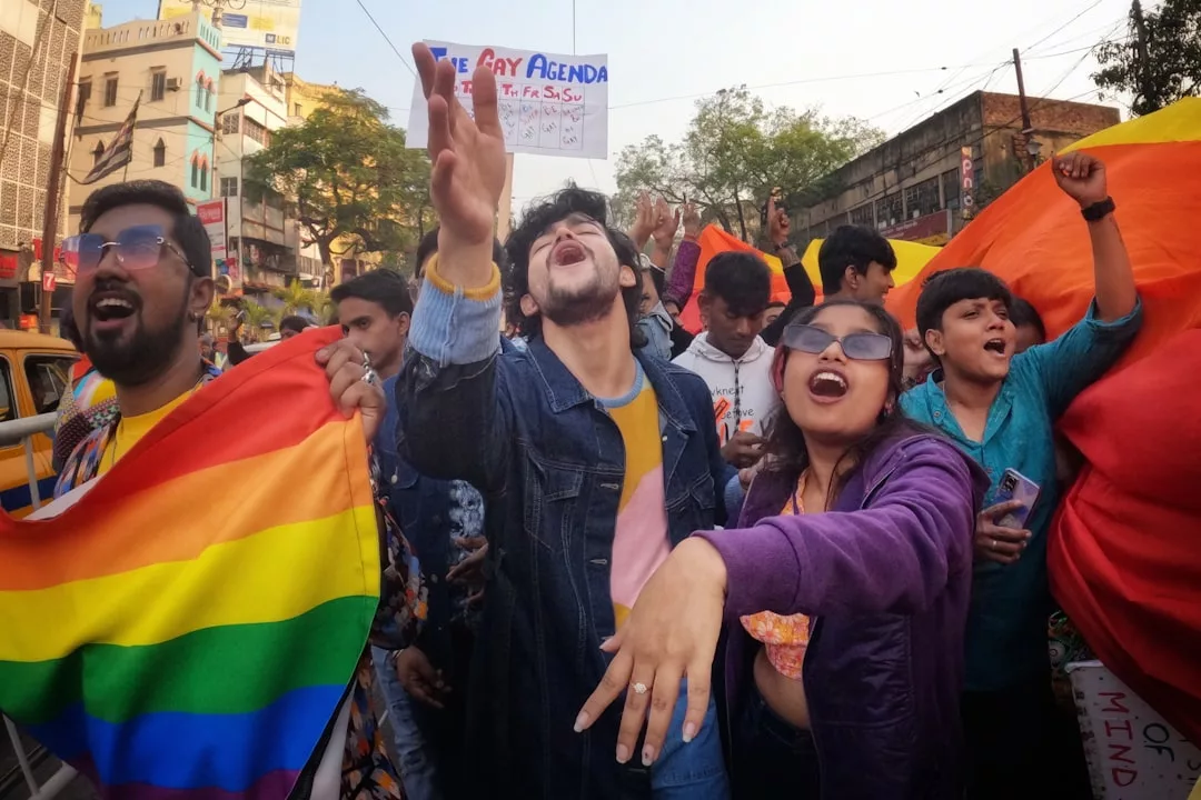 A group of people holding rainbow flags participate in a pride parade on a city street, expressing enthusiasm and solidarity as local real estate businesses show their support for the LGBTQ+ community.