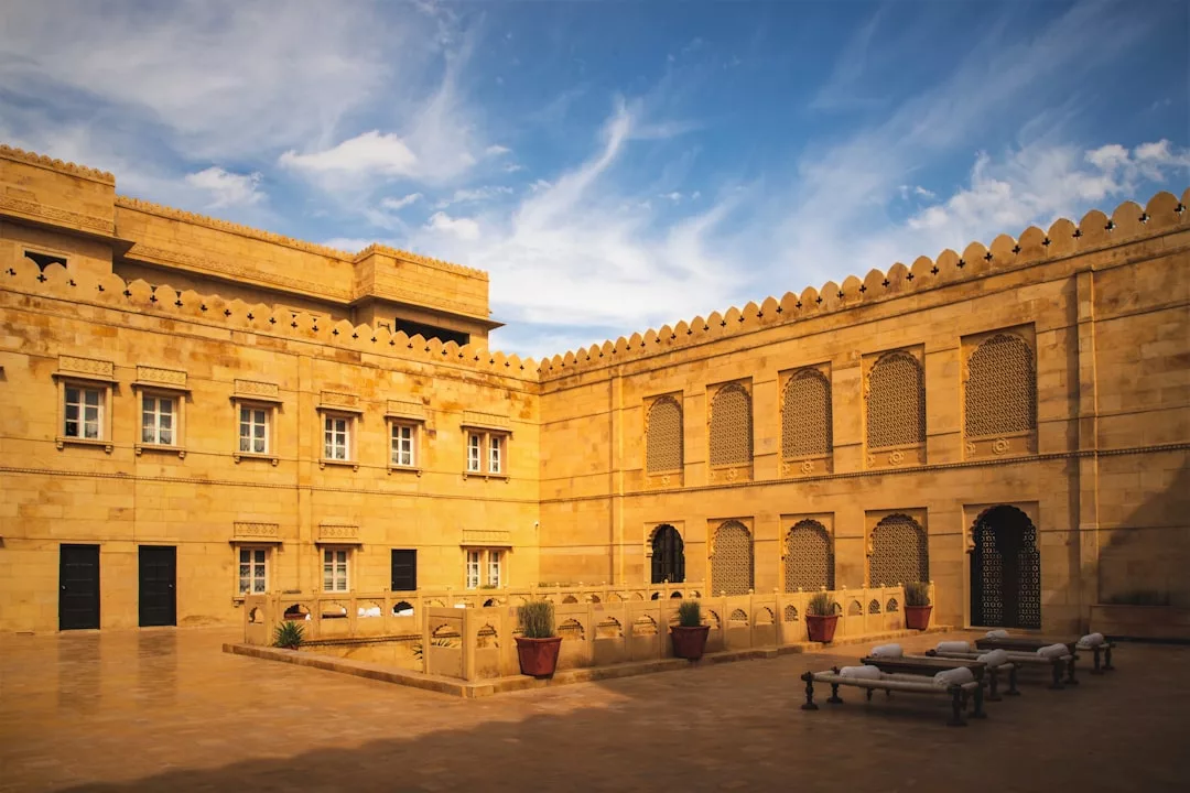 A real estate gem, this courtyard features sand-colored stone walls, arched windows, and decorative latticework, with benches and potted plants beneath a blue sky adorned with wispy clouds.