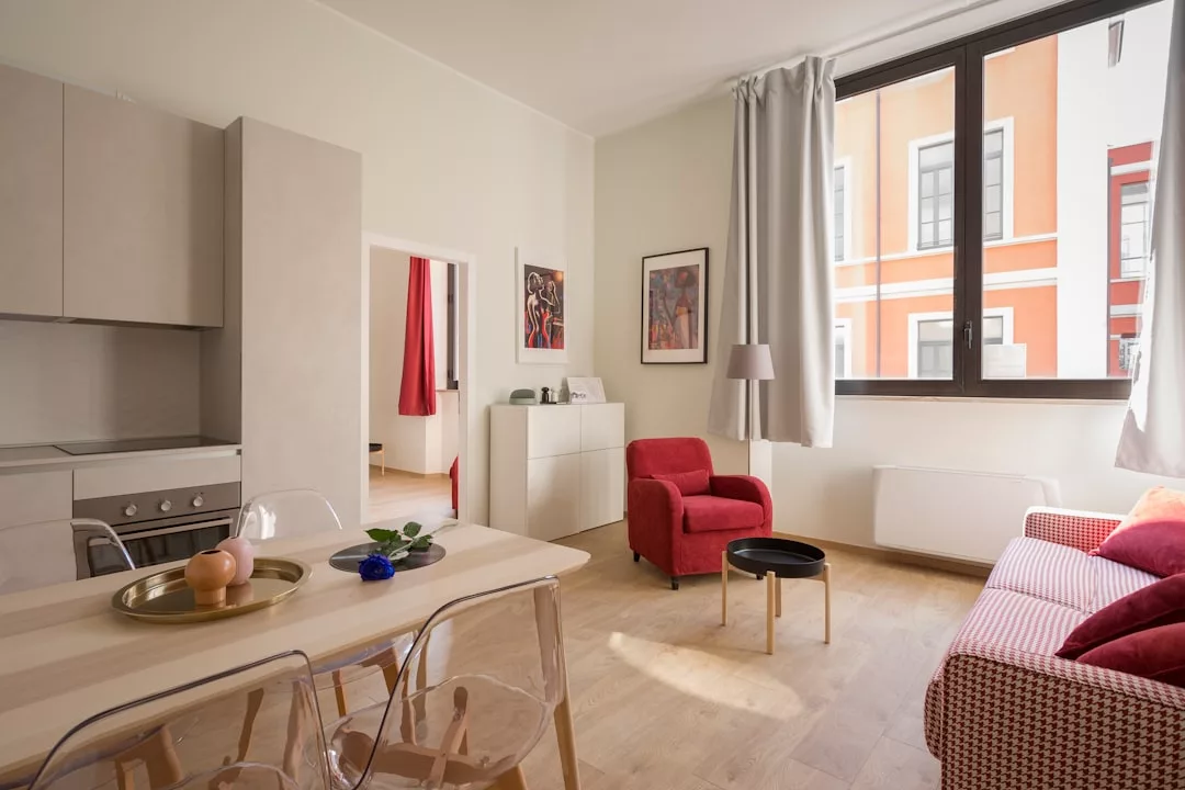 Modern living room ideal for real estate buyers, featuring light wood floors, red armchair, sofa, small table, dining area, and large window with natural light. Kitchen with oven is visible on the left.