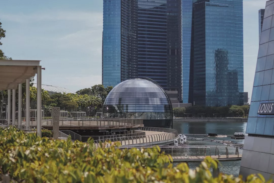 A glass dome building sits on the water in front of tall modern skyscrapers, showcasing premium real estate, with lush greenery and a Louis Vuitton sign in the foreground.