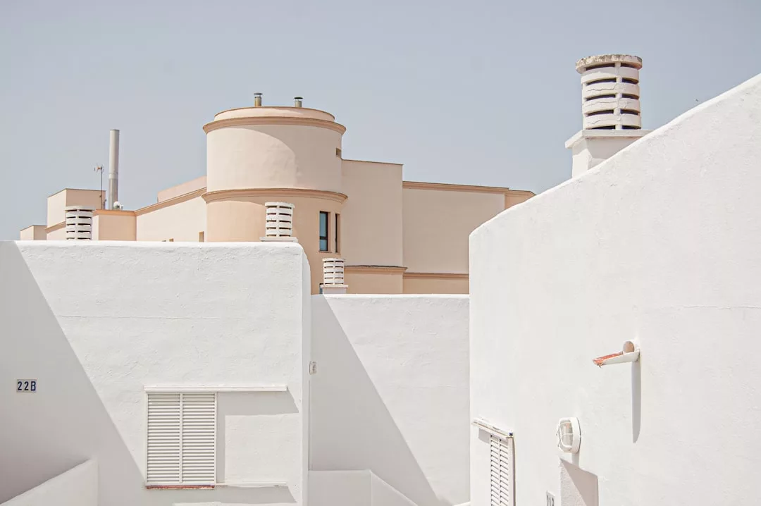 White-walled buildings with cylindrical chimneys and a beige, rounded structure in the background stand under a clear sky, offering unique real estate charm.