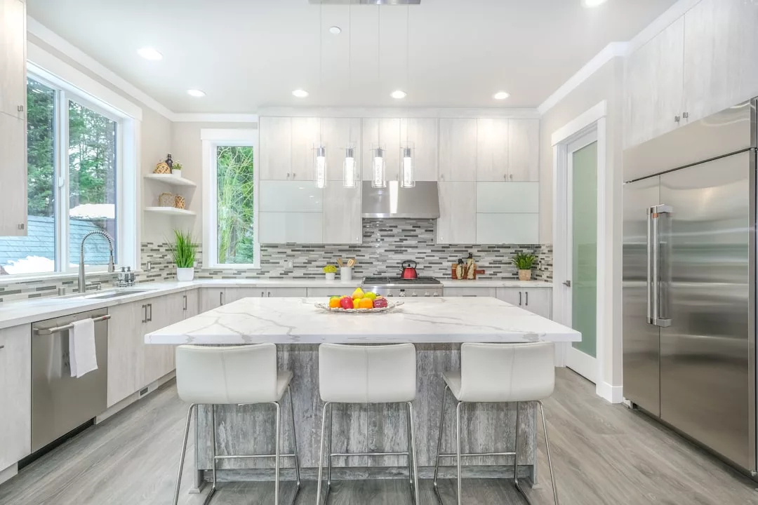 Modern kitchen in a real estate gem with a large marble island, four white stools, stainless steel appliances, sleek white cabinets, and a window overlooking greenery. Fruit bowl sits on the island.