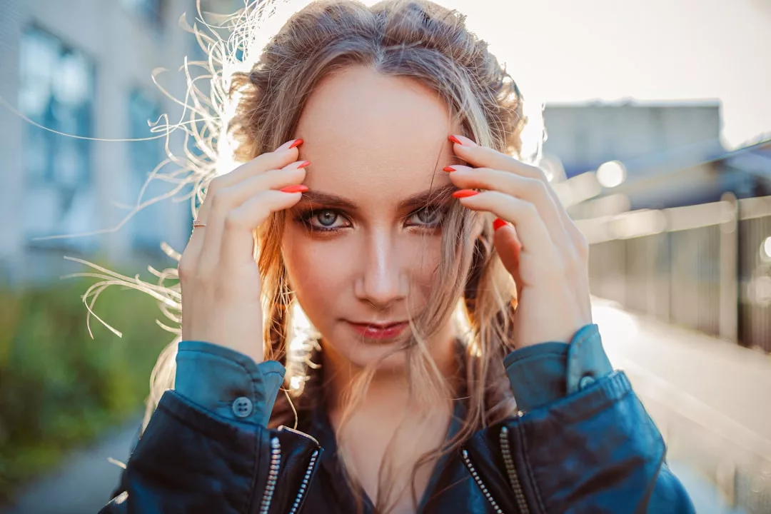 A woman with long hair and painted nails gazes confidently at the camera, her hands near her face, outdoors with sunlight in the background—capturing elegance perfect for a real estate promotional shoot.