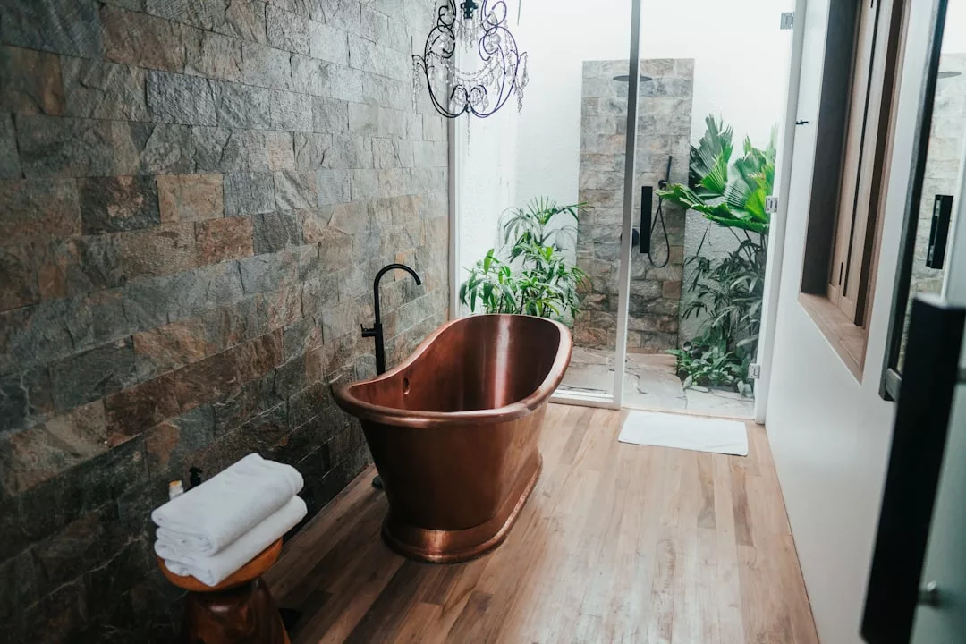 A stunning real estate bathroom features a copper bathtub, a folded towel stack on a stool, stone accent walls, wood flooring, a glass door, and lush plants visible outside.
