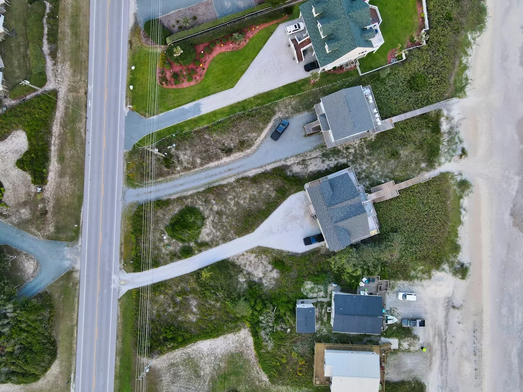 Aerial view of three real estate properties near a road with driveways, greenery, and sand patches, bordered by a beach area on the right side.