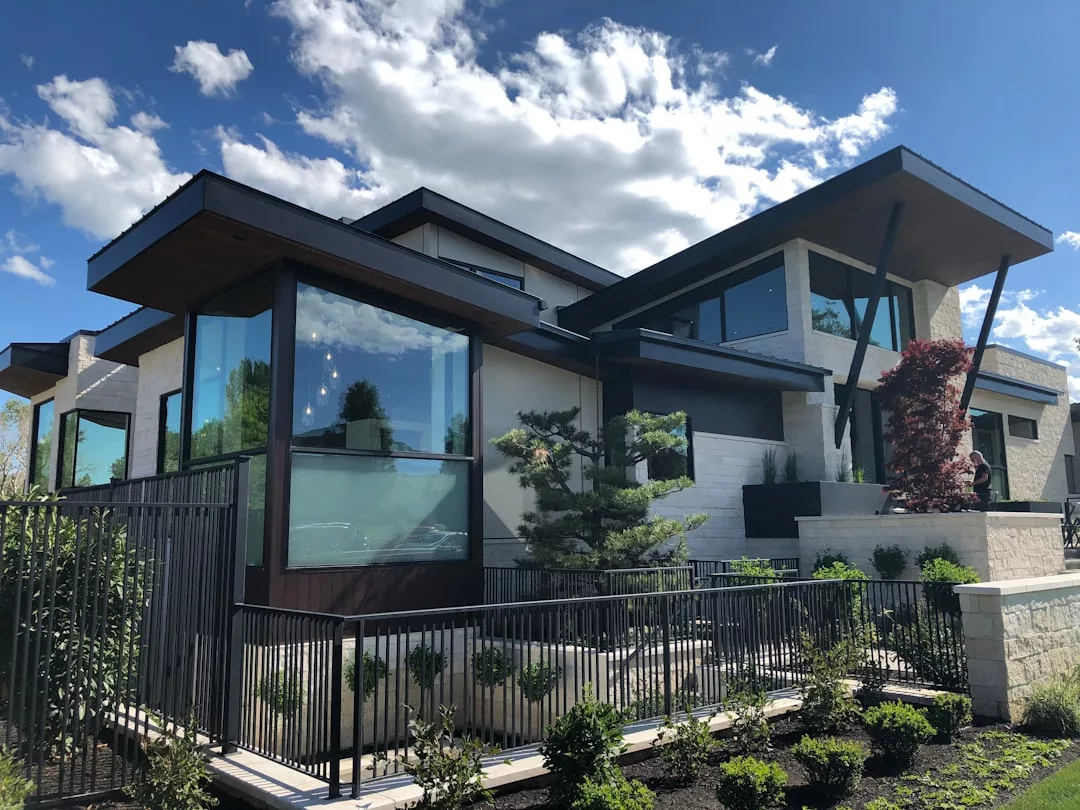 Modern two-story real estate property with large glass windows, flat roofs, a metal railing, and a landscaped front yard under a partly cloudy sky.