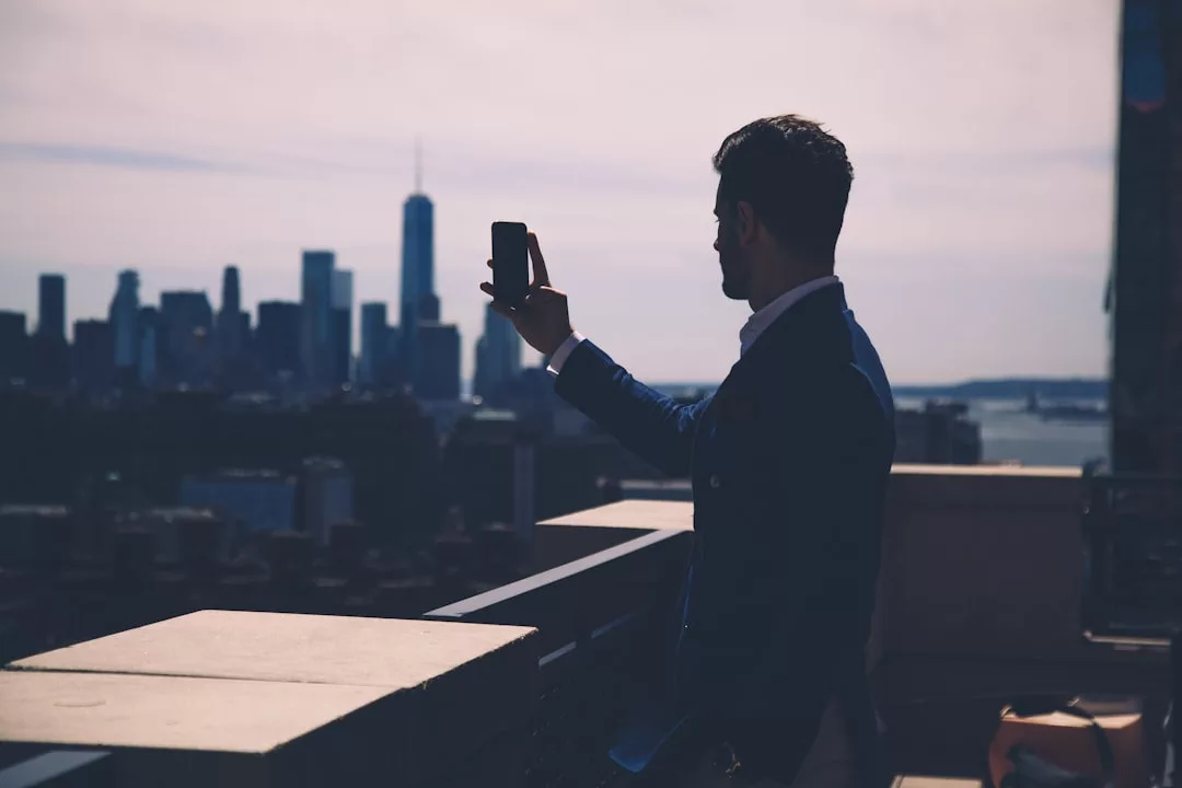 A man in a suit stands on a balcony taking a selfie with his smartphone, capturing the city skyline and tall buildings—a perfect moment for any real estate professional.