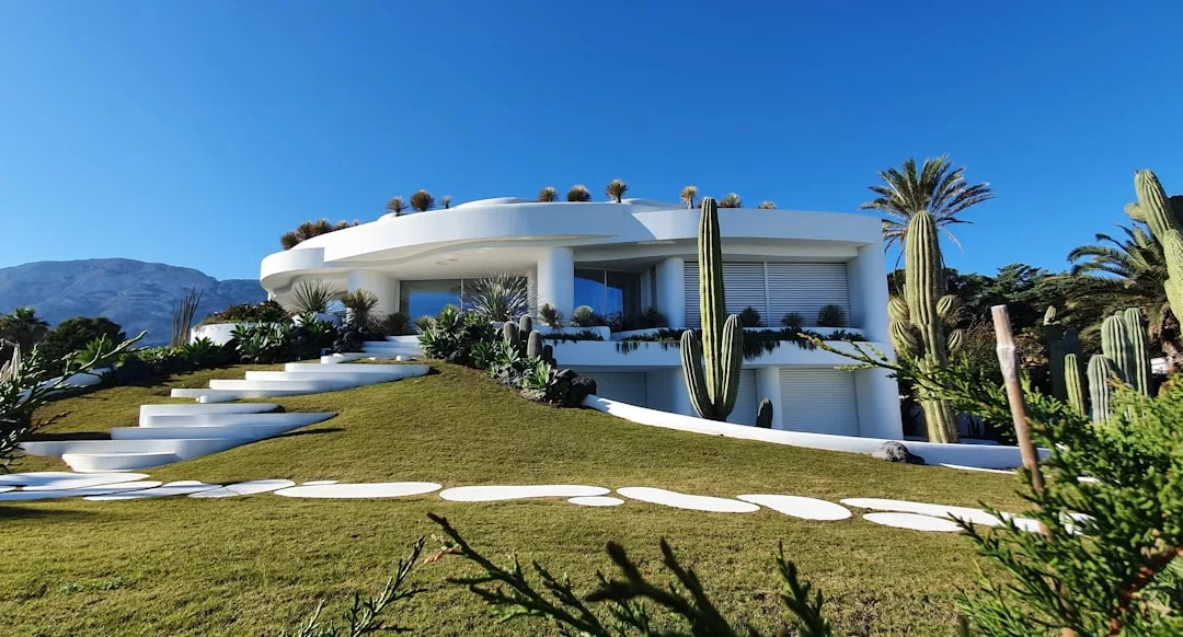 Modern white two-story house with large windows, surrounded by cacti and palm trees, on a grassy hill with a curved stone pathway, under a clear blue sky—an ideal real estate opportunity.