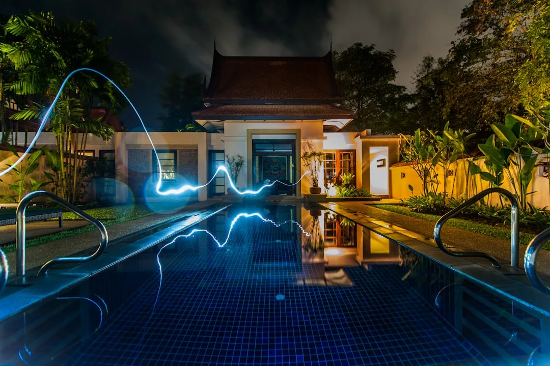 Night view of a house with a traditional roof, a lit swimming pool in the foreground, and a blue light streak above the water—perfect real estate surrounded by lush greenery.