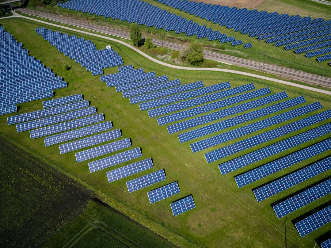 Aerial view of a large solar farm with rows of blue solar panels on green grass, intersected by a road and railway tracks—an innovative real estate development blending sustainable energy with modern infrastructure.