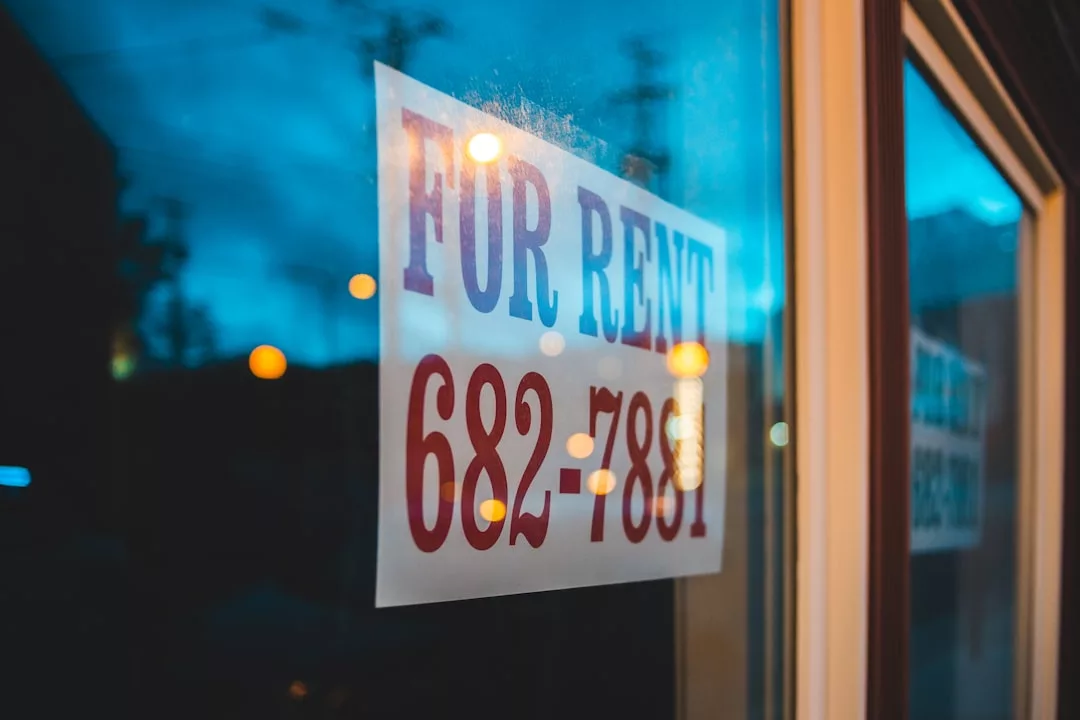 A "For Rent" sign with a phone number is displayed on a window, photographed during dusk with lights reflected in the glass—a classic image of real estate opportunities waiting to be discovered.