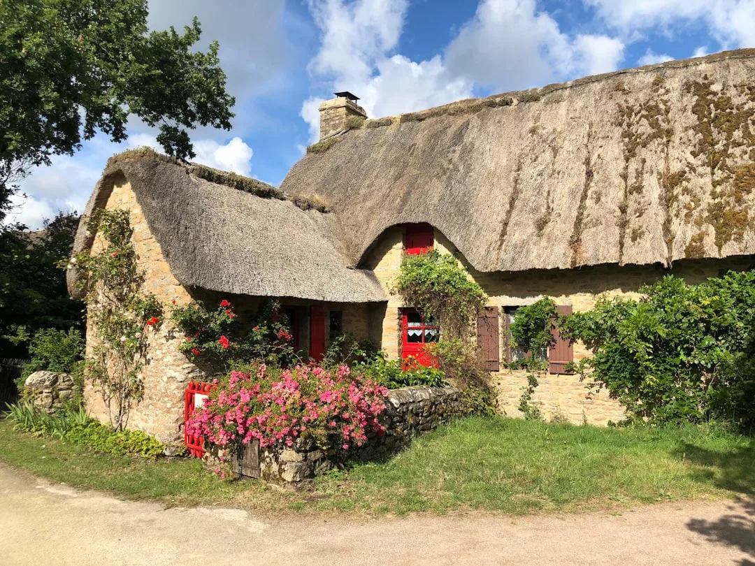 Charming stone cottage with a thatched roof, red window frames, and flowers climbing around the entrance—an inviting real estate gem set against a backdrop of trees and blue sky.