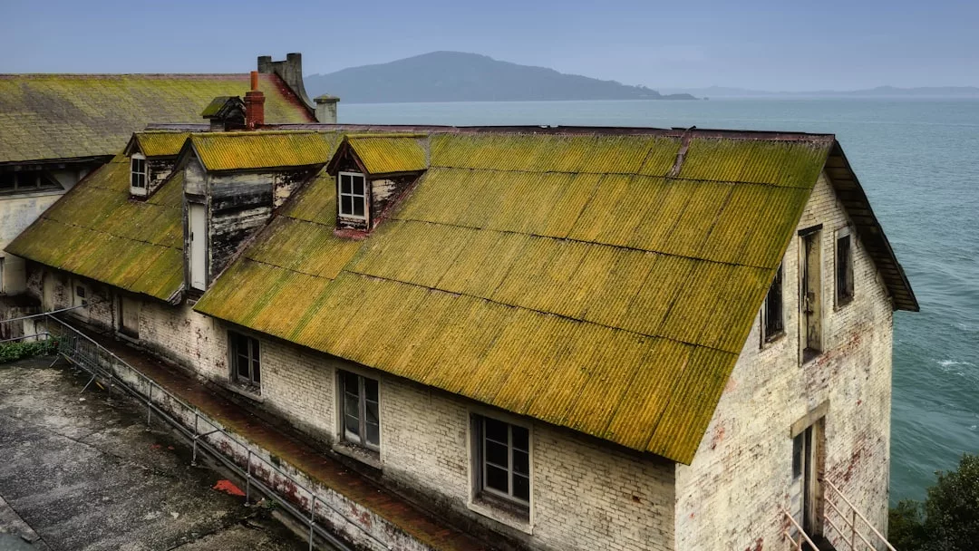A large, weathered real estate building with yellow-green moss on the roof sits by the edge of a body of water, with hills visible in the background.