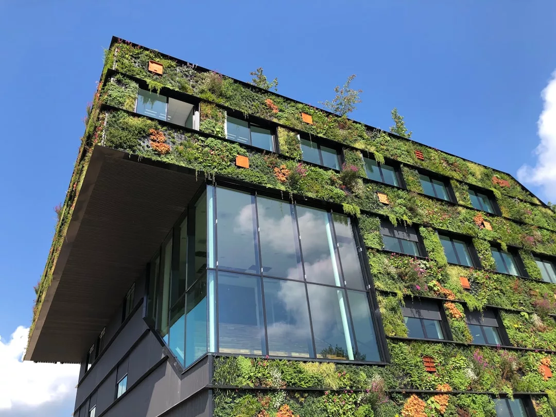 A modern real estate building with large windows and a green facade covered in plants, set against a clear blue sky with some clouds.