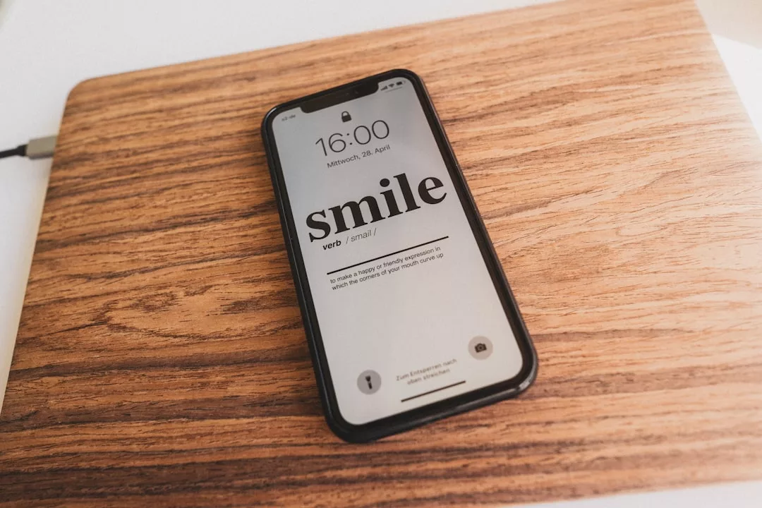 A smartphone on a wooden surface displays a lock screen with the time 16:00 and the word "smile" defined as a verb, reflecting positivity ideal for real estate professionals connecting with clients.
