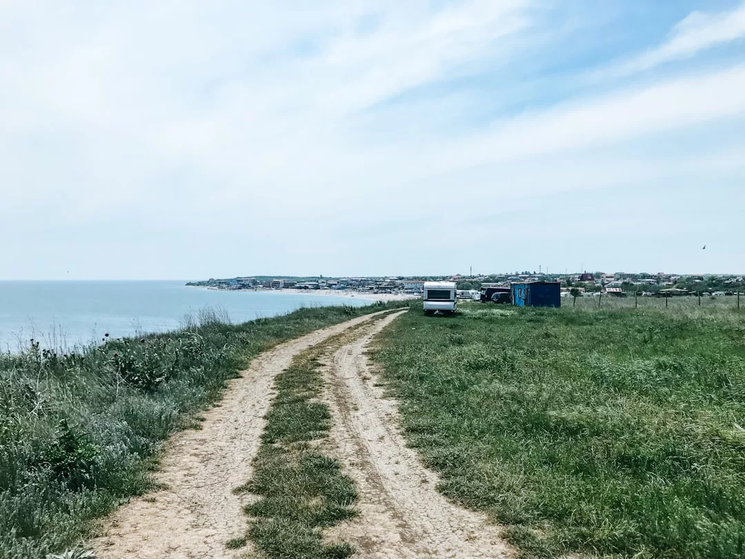 A dirt road curves along a grassy field by the sea, leading toward a distant town. Perfect for real estate opportunities, a white van and blue structure are parked near the road under a partly cloudy sky.