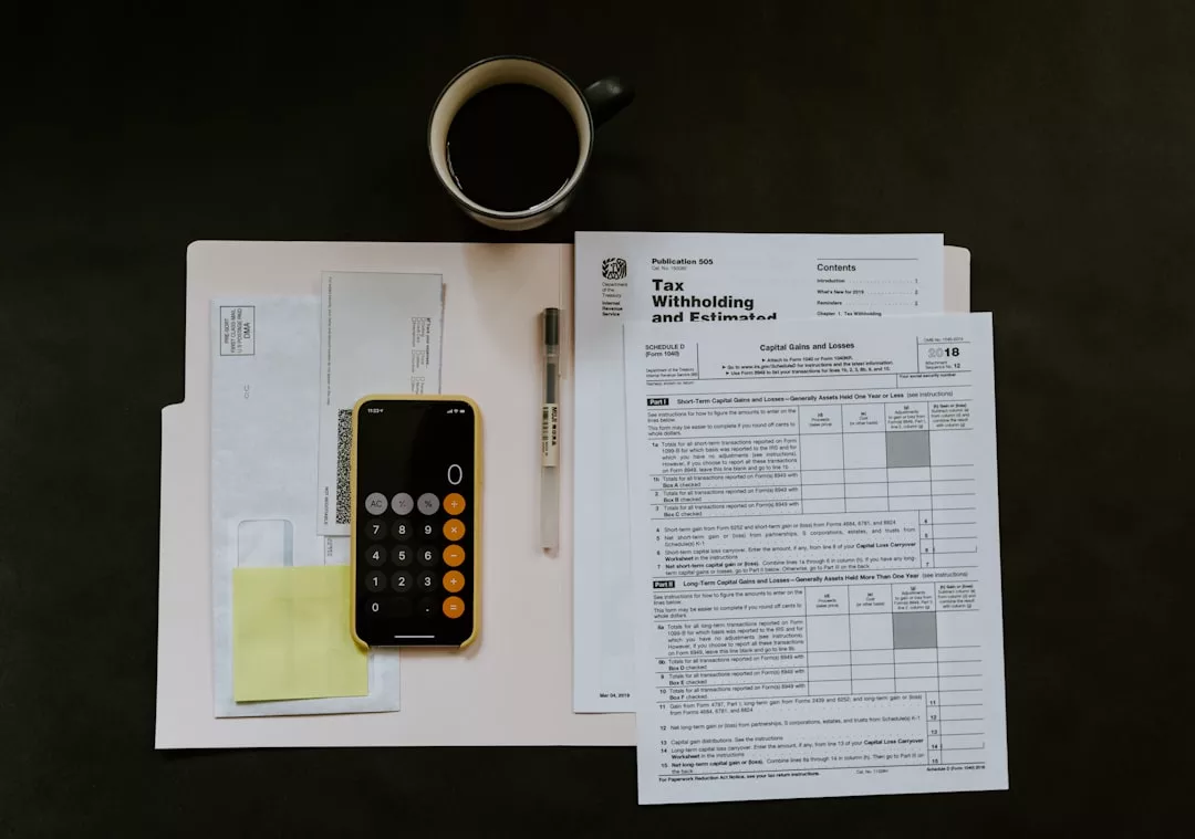 A tax folder with real estate documents, IRS forms, a smartphone displaying a calculator, a pen, sticky notes, and a cup of black coffee on a dark surface.