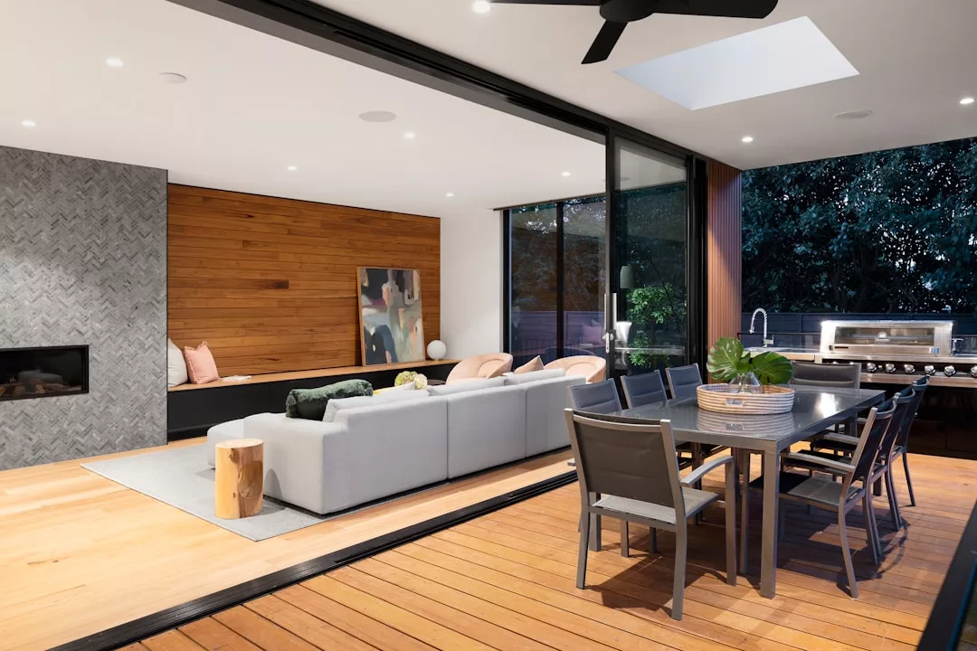 Modern open-plan living and dining area in this stunning real estate listing features a gray sectional sofa, wooden accent wall, outdoor dining table, and stainless steel grill on a spacious wooden deck.
