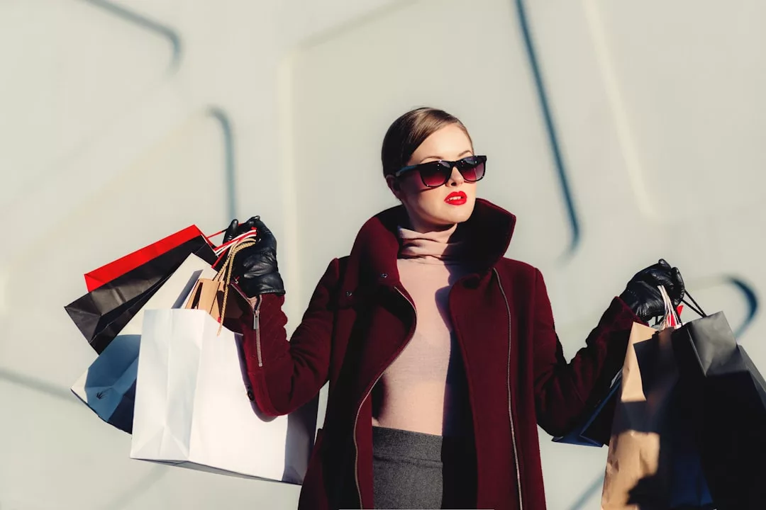 Woman in sunglasses and a burgundy coat holds multiple shopping bags in both hands, standing confidently as if she just closed a Real Estate deal, against a light-colored wall.