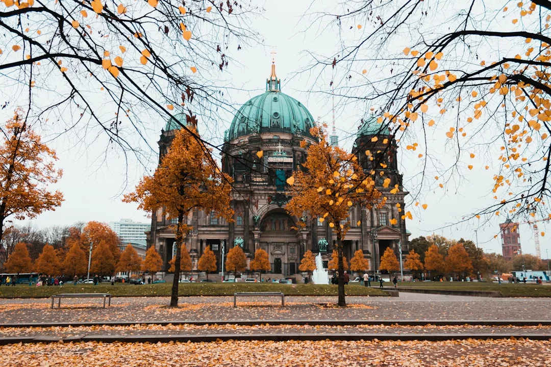 Berlin Cathedral with autumn trees and fallen leaves in the foreground, cloudy sky above, and benches on the lawn—an iconic location admired by real estate enthusiasts for its picturesque charm and vibrant city atmosphere.