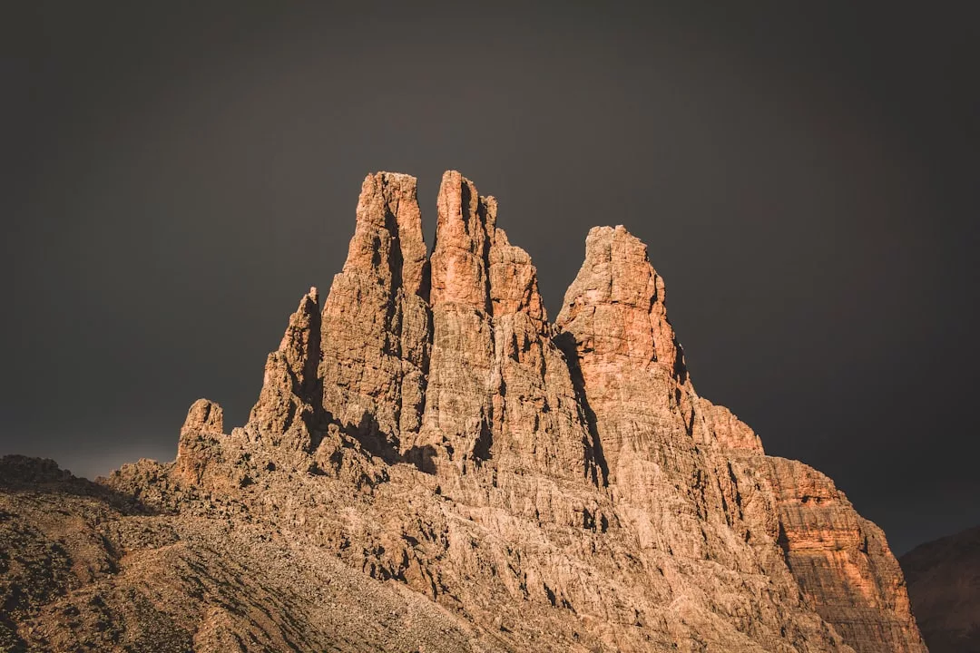 Rocky mountain peaks rise sharply against a dark, overcast sky, with sunlight illuminating the rugged rock formations—an inspiring backdrop for premium real estate nestled in nature’s grandeur.