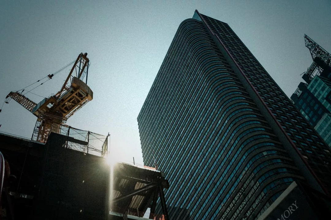 A modern high-rise building and a construction crane stand tall against a clear sky, with sunlight peeking through the construction site, capturing the dynamic growth of real estate development.