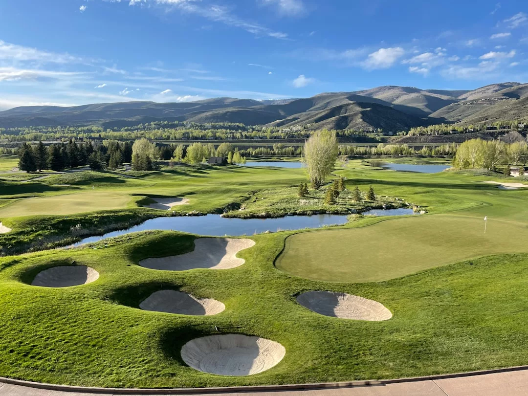 A stunning golf course with multiple sand bunkers, green fairways, small ponds, and mountain views—perfect for real estate enthusiasts seeking scenic surroundings under a clear blue sky.