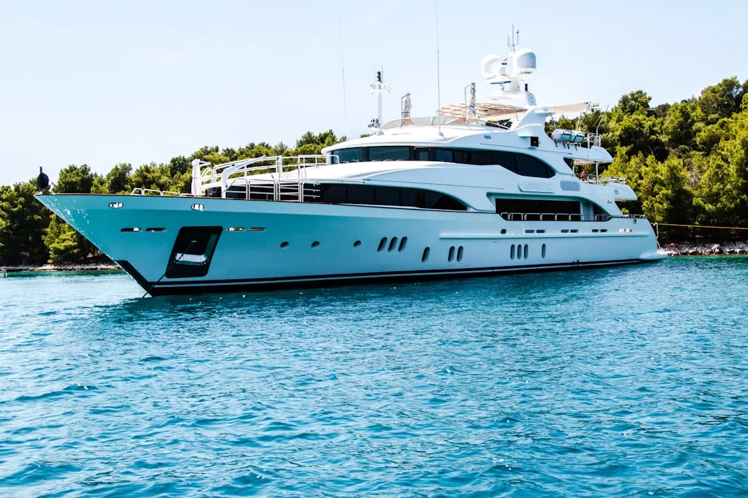 A large white luxury yacht, reminiscent of elite real estate, is anchored on calm blue water near a forested shoreline under a clear sky.