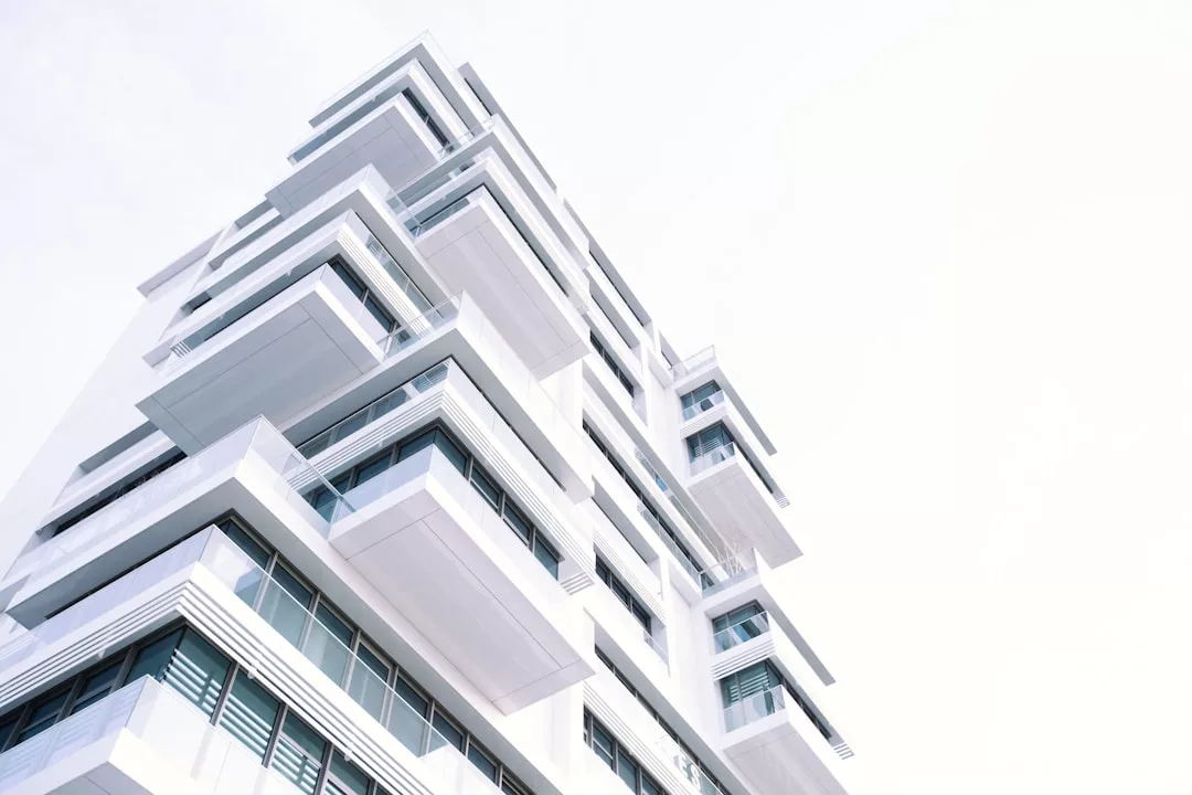 Modern white apartment building with multiple large balconies and glass windows, viewed from a low angle against a bright, overexposed sky—an impressive example of contemporary real estate.