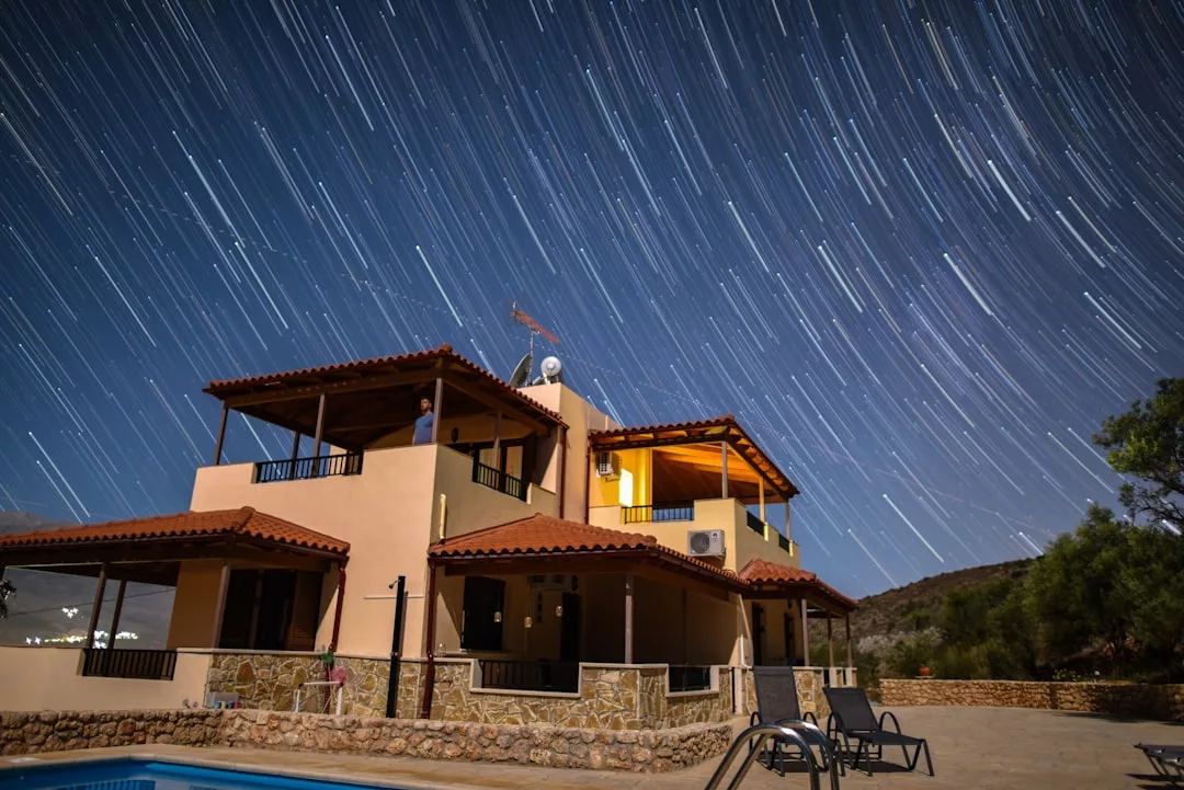 A stunning real estate gem, this two-story house with a red-tiled roof is showcased at night under star trails, featuring a sparkling swimming pool and inviting lounge chairs in the foreground.