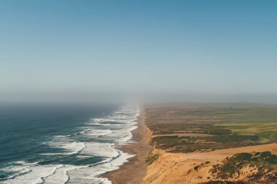 A wide view of a coastline with waves crashing on a sandy beach, exposed cliffs, and green fields stretching inland under a clear blue sky—an ideal real estate opportunity for those seeking natural beauty and serenity.