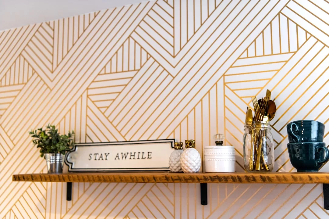 A wooden shelf in this inviting real estate space holds a potted plant, a "Stay Awhile" sign, decorative jars, utensils in a glass, and stacked blue cups against a gold and white geometric patterned wall.