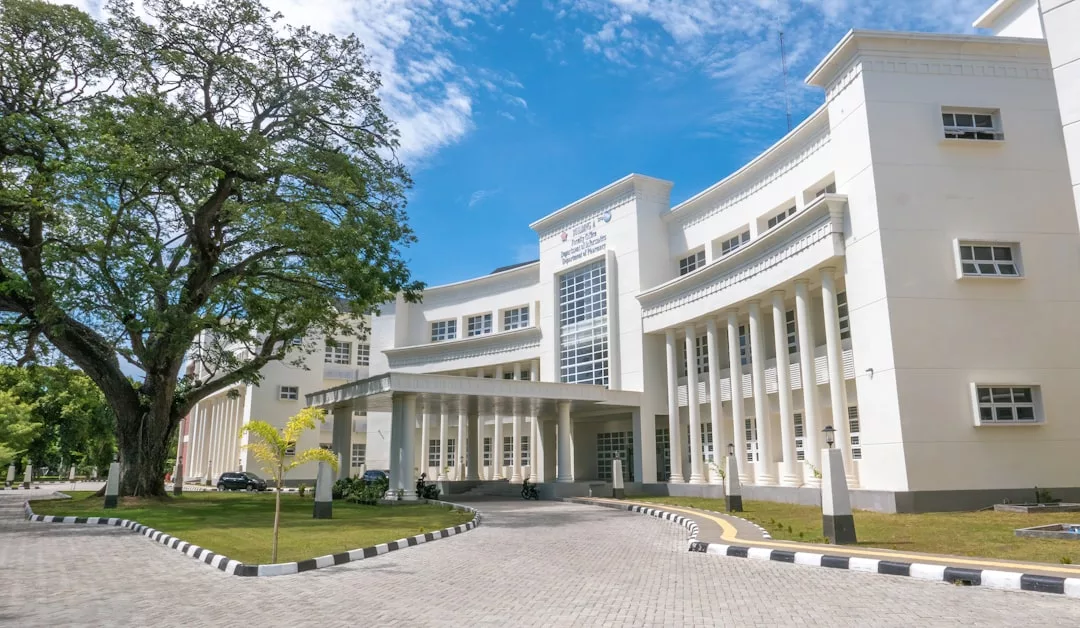 A large, modern white real estate building with tall columns and a glass entrance stands next to a leafy tree under a partly cloudy sky.