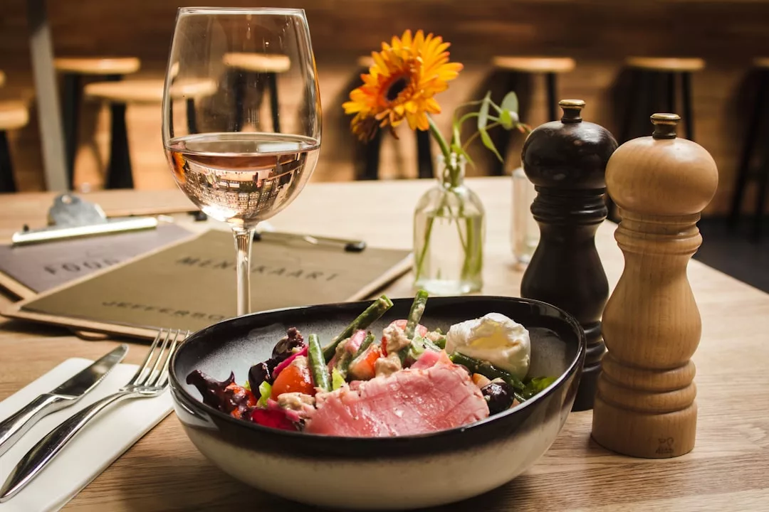 A bowl of salad topped with seared tuna and cheese sits on a table, styled like a luxury real estate setting, with rosé wine, salt and pepper shakers, cutlery, and a small vase with a yellow flower.