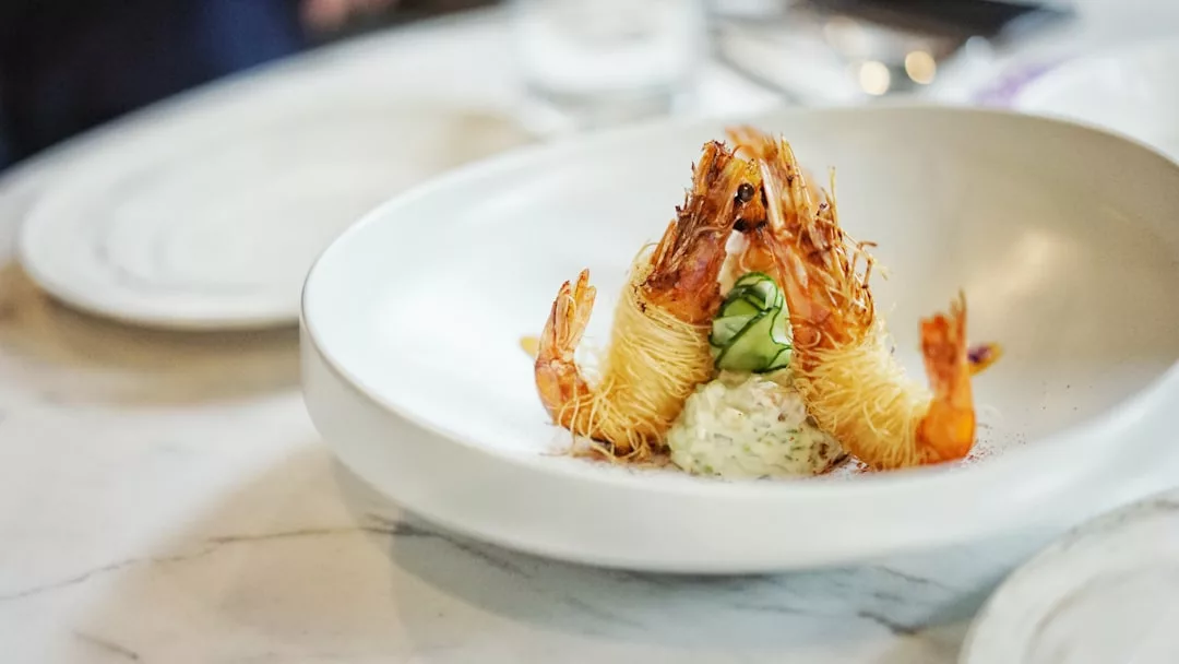 A white plate with three kataifi-wrapped shrimp arranged upright, served with a dollop of creamy sauce and cucumber slices on a marble table—an elegant touch reminiscent of upscale real estate dining experiences.