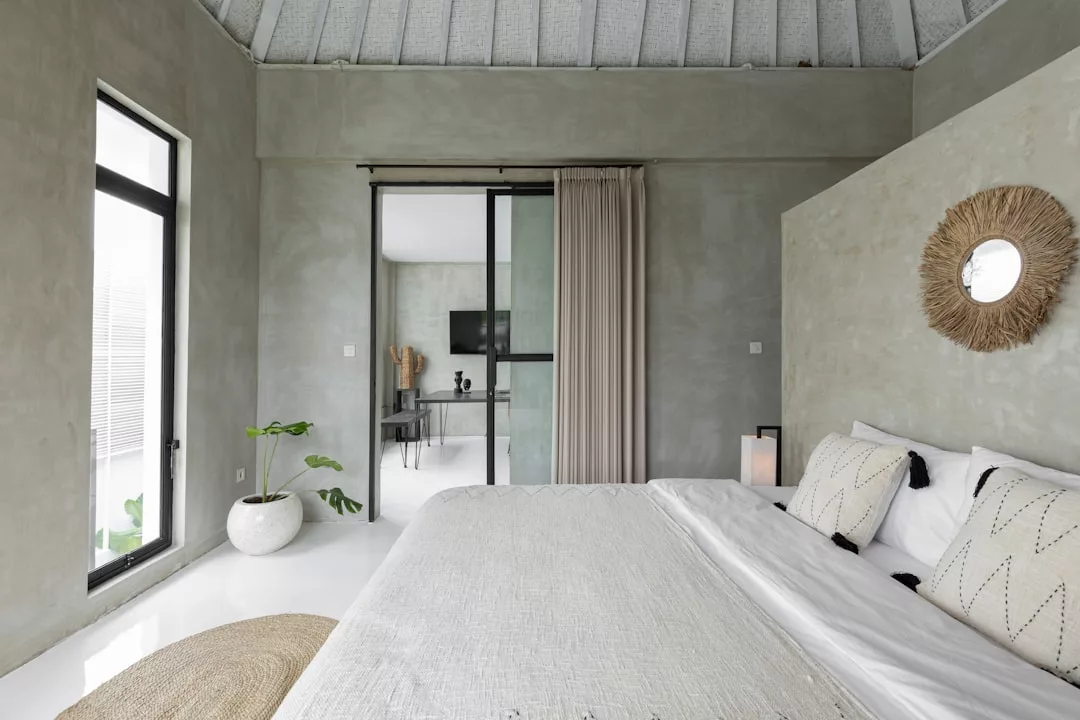Minimalist bedroom with a neatly made bed, neutral tones, decorative pillows, wall mirror, large windows, potted plant, and sliding glass doors—perfect for showcasing elegant real estate.