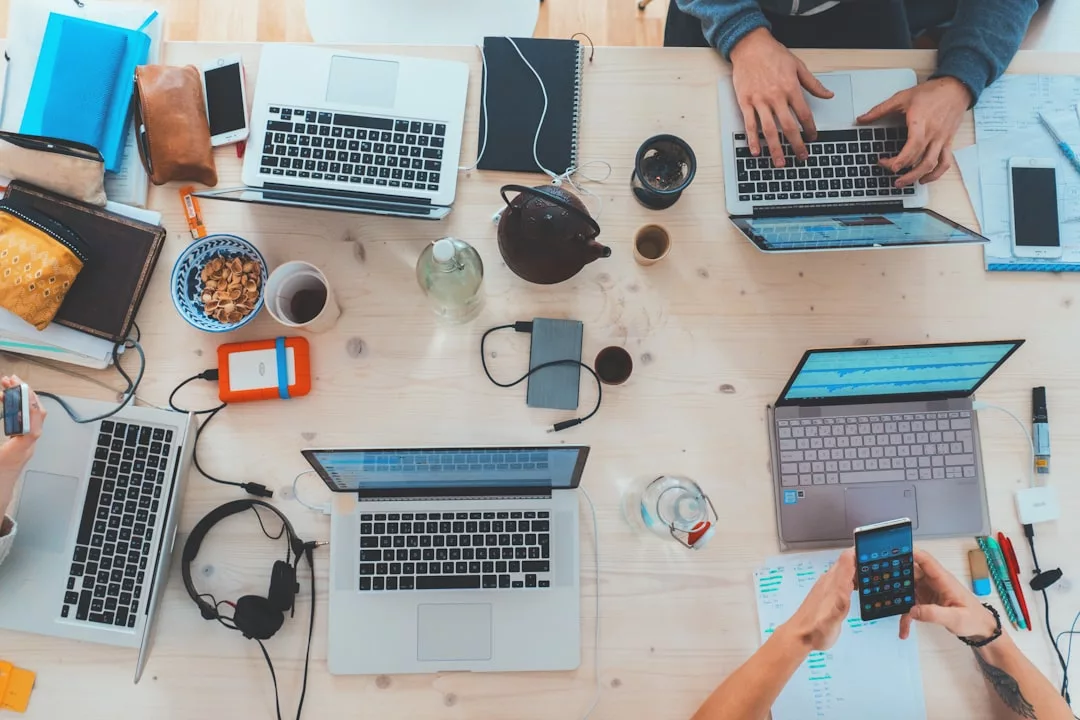Four people work at a cluttered desk with laptops, notebooks, snacks, headphones, and smartphones—perhaps collaborating on a Real Estate project—viewed from above.