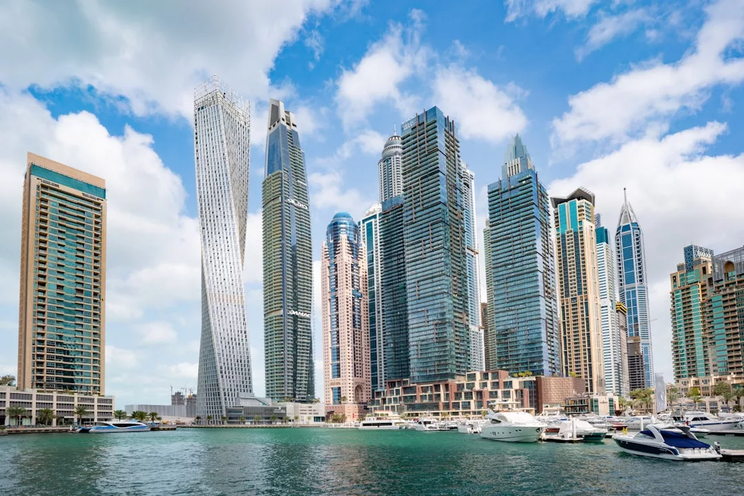 Modern skyscrapers line the waterfront in a city marina, highlighting prime real estate, with several docked yachts and boats visible in the foreground under a partly cloudy sky.