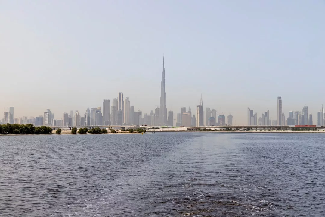 A city skyline with numerous high-rise buildings and a tall central tower, showcasing prime real estate opportunities, is seen from across a body of water under a clear sky.