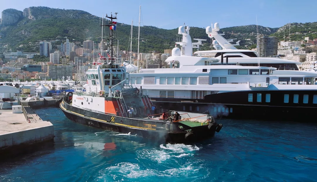 A tugboat maneuvers near a large white yacht in a marina, set against buildings and green hills—an inviting waterside scene that highlights the appeal of local real estate.