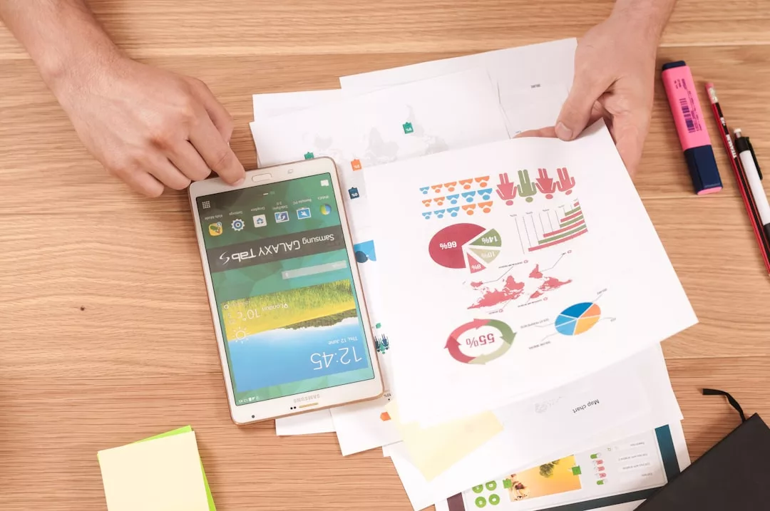 A person holds a sheet with colorful real estate charts and graphs above a stack of papers, next to an upside-down Samsung tablet, pens, and highlighters on a wooden desk.