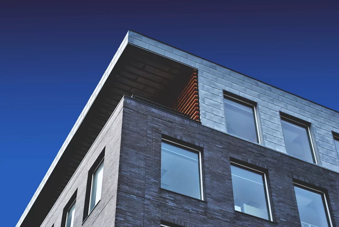 A modern real estate building with a gray brick and metal facade, large windows, and a covered corner section, viewed from below against a clear blue sky.