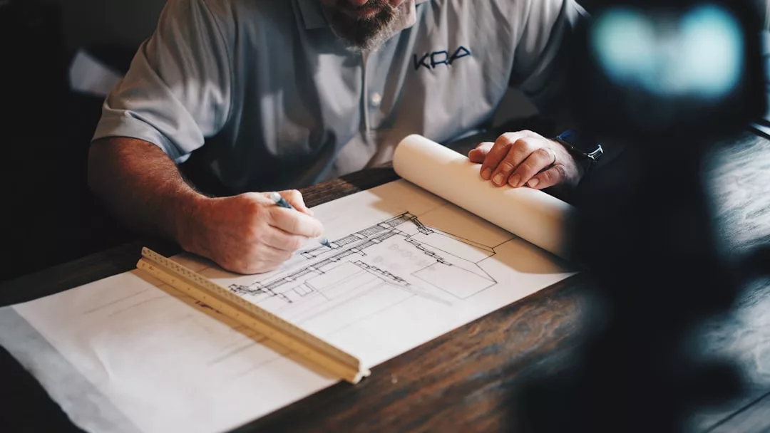 A person in a collared shirt sketches architectural plans on a large sheet of paper at a desk, with a ruler and rolled-up real estate plans nearby.