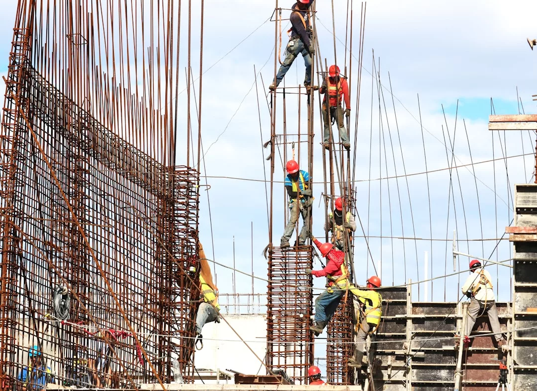 Construction workers wearing safety gear are working on a real estate building site, assembling steel reinforcement bars for concrete structures under a clear sky.