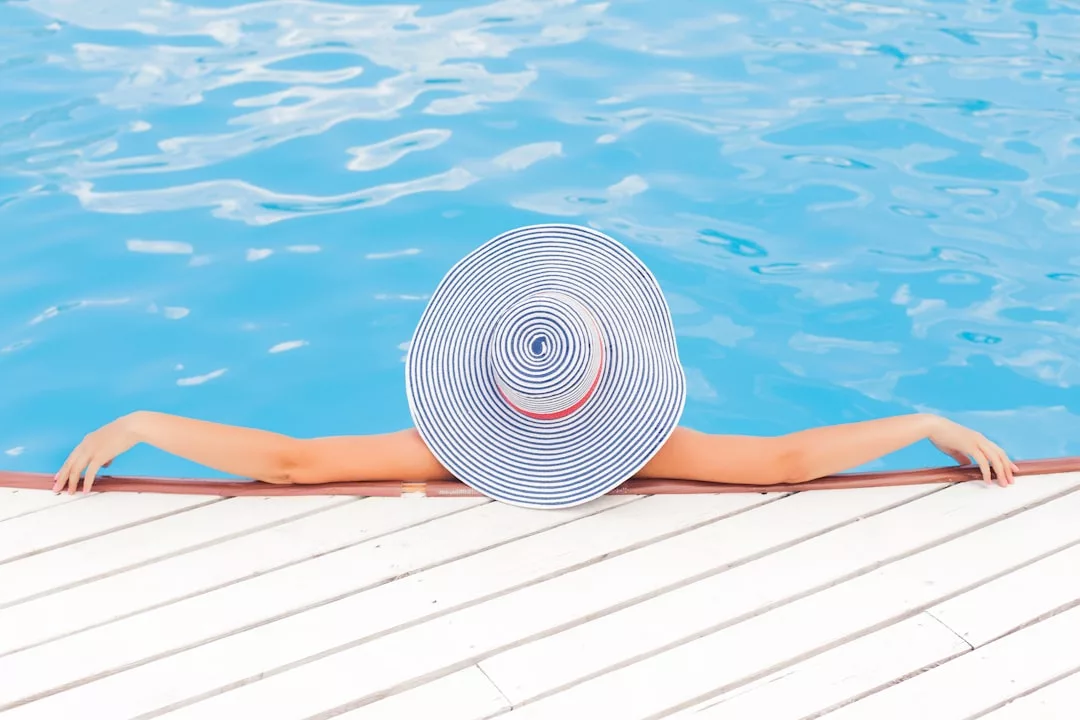 A person in a wide-brimmed striped hat relaxes in a swimming pool, arms resting on the edge of a white wooden deck—a perfect scene showcasing luxury real estate living.