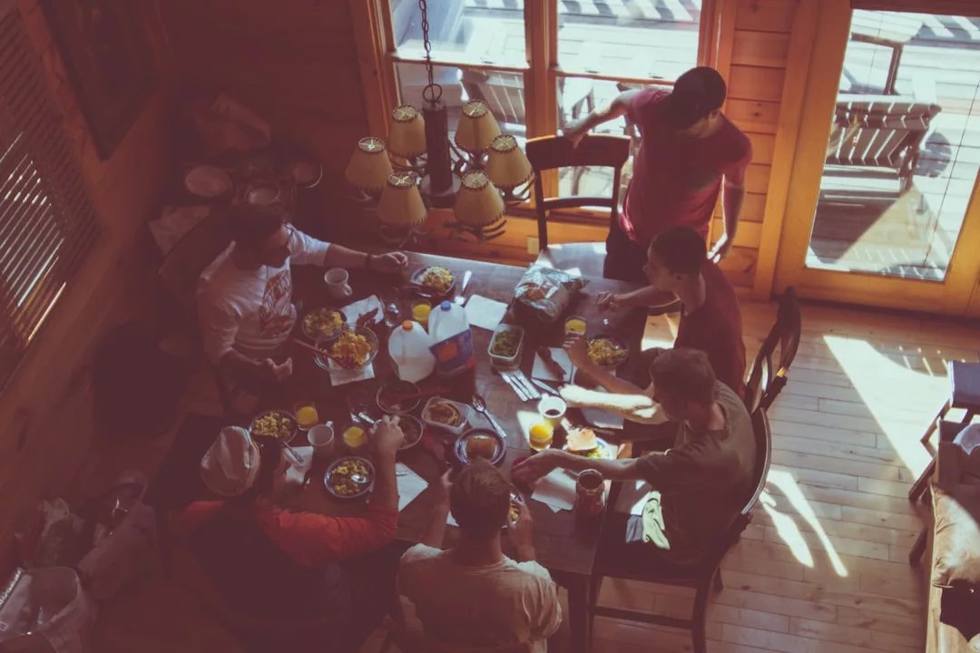 Six people sit around a wooden dining table having a meal together in a sunlit room with large windows, showcasing the inviting atmosphere of this beautiful real estate.