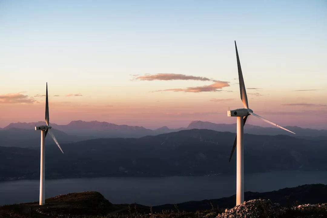 Two wind turbines stand on a hill at sunset, overlooking a body of water and mountains—an inspiring view that highlights the appeal of sustainable real estate in scenic locations.