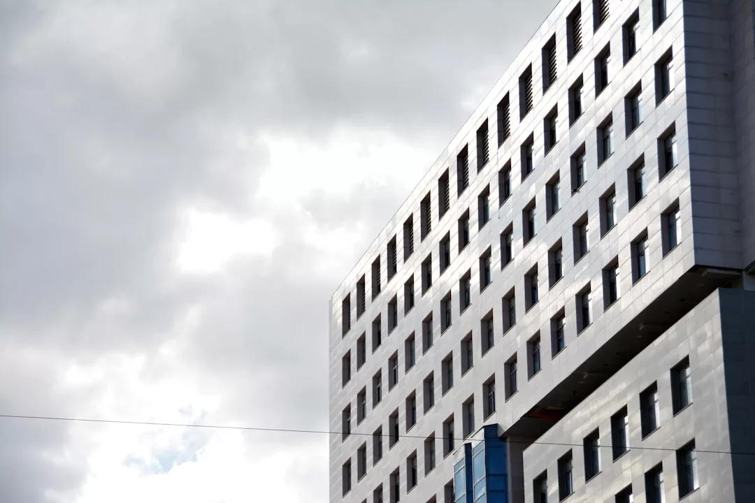 A multi-story modern real estate building with a grid of rectangular windows, photographed from below against a cloudy sky.