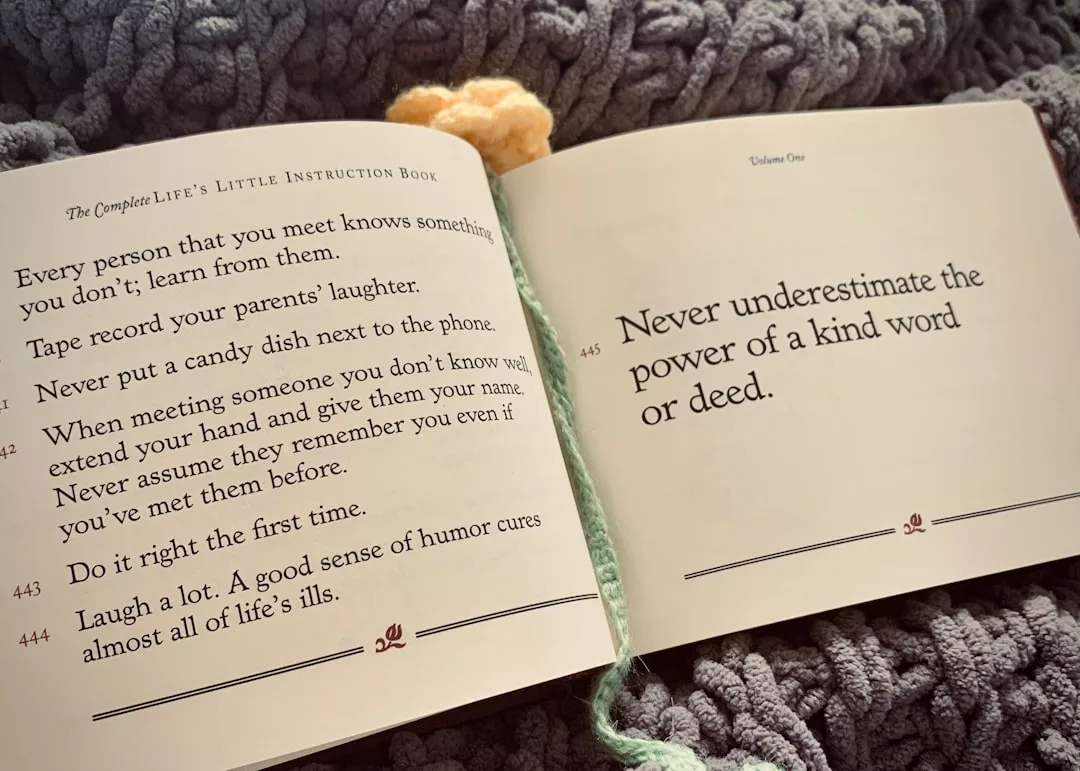An open book lies on a chunky grey knit blanket, displaying life advice tips—like “Never underestimate the power of a kind word or deed.” Nearby, a yellow crocheted flower adds warmth, creating a cozy scene perfect for real estate inspiration.