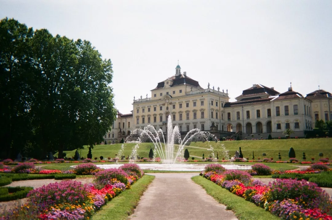 A large baroque palace with a central fountain in front, surrounded by colorful flower gardens and neatly trimmed lawns on a sunny day—a true showcase of luxurious real estate.